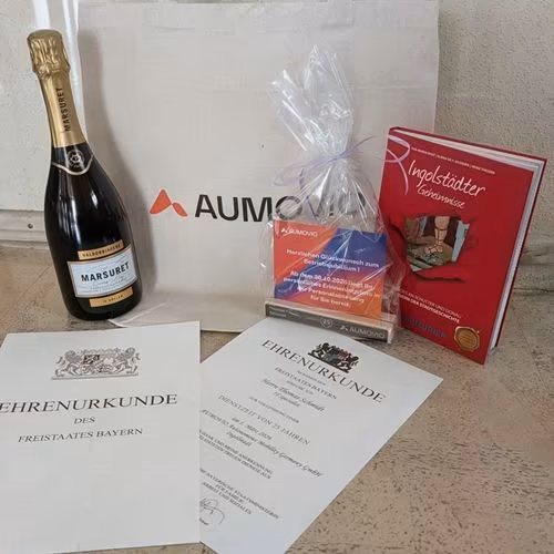 Stillleben zur Feier eines 25-jährigen Firmenjubiläums in Ingolstadt. Zu sehen sind eine Ehrenurkunde des Freistaates Bayern, das Buch „Ingolstädter Geheimnisse“, eine Flasche Sekt, ein AUMOVIO-Jubiläumspräsent zum 25-jährigen Dienstjubiläum sowie eine AU