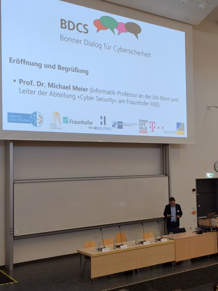 Ein Mann steht auf einer Bühne mit Tisch und Stühlen. Hinter ihm auf einer Leinwand steht „Bonner Dialog für Cybersicherheit“ und dass es sich um Prof. Dr. Michael Meier handelt, der die Anwesenden begrüßt.
