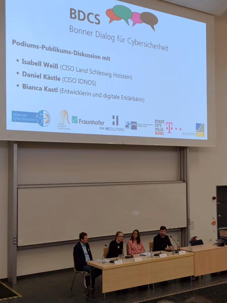 Vier Personen sitzen an einem Tisch mit Mikrofonen. Hinter ihnen auf einer Leinwand stehen ihre Namen: Isabell Weiß (CISO Land Schleswig Holstein), Daniel Kästle (CISO IONOS), Bianca Kastl (Entwicklerin und digitale Erklärbärin)